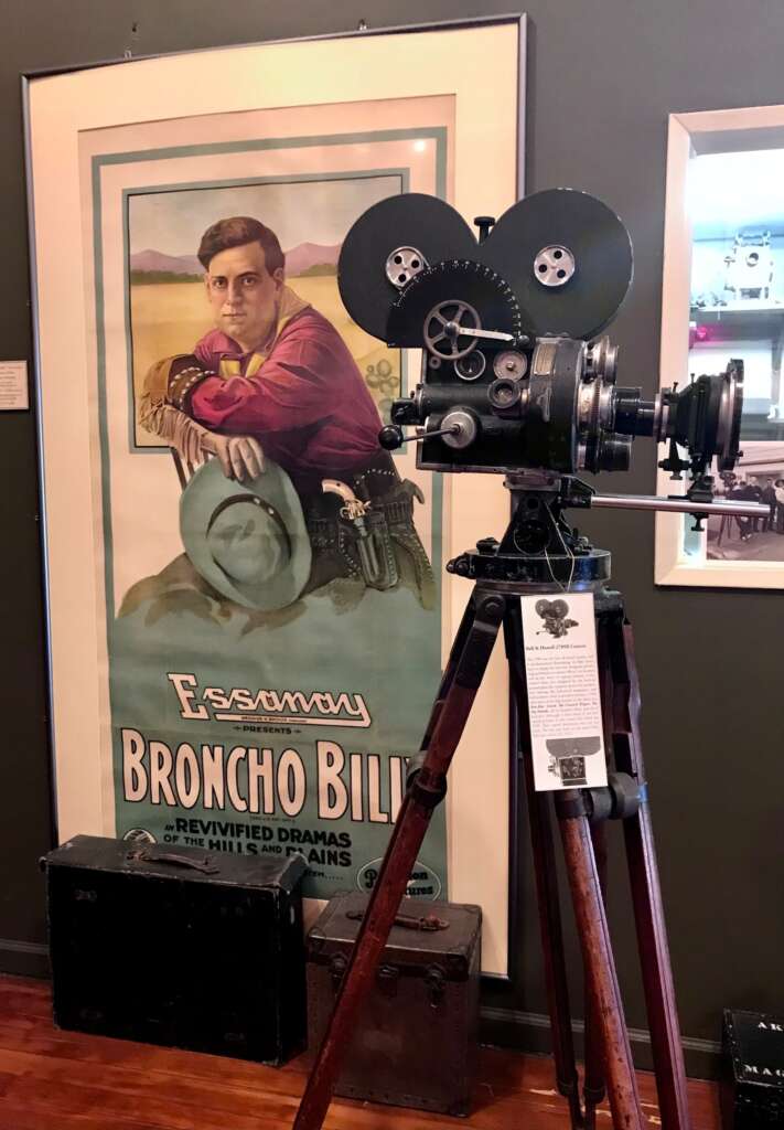 Photo shows an old movie poster with a silent film camera in front of it.