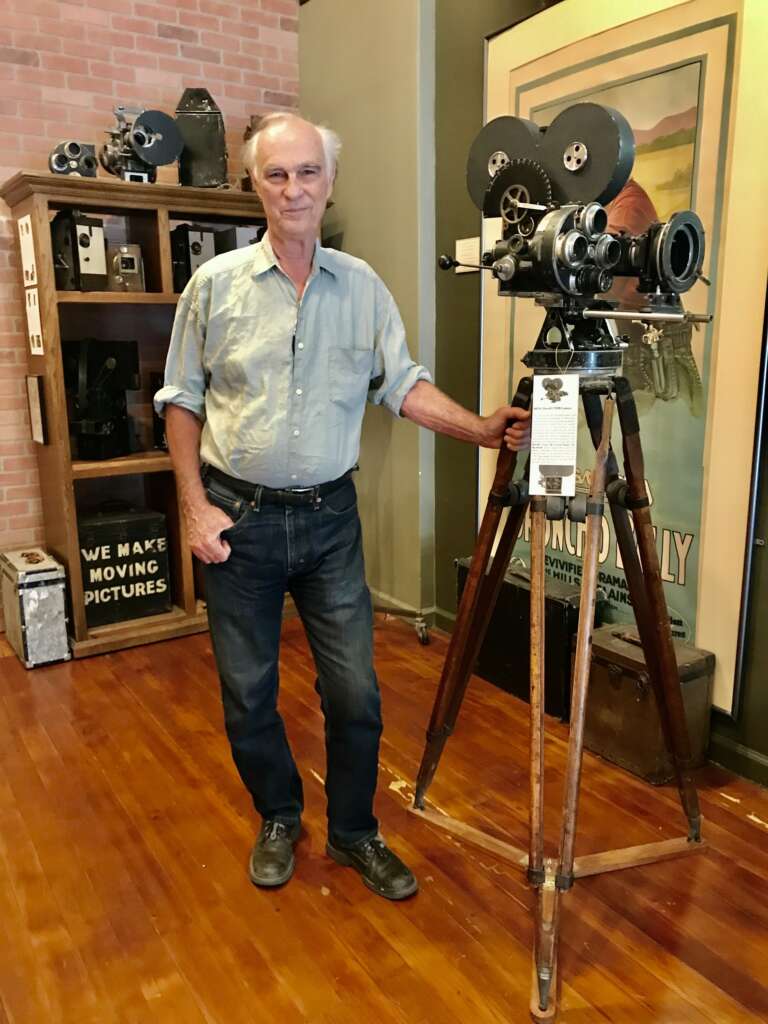 An older man standing next to a silent film camera with museum shelves behind.