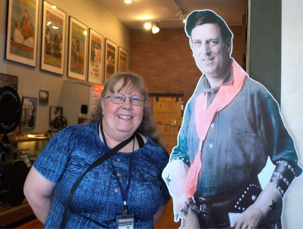 A woman in blue standing next to a large cardboard cutout of a silent film star with museum displays behind.